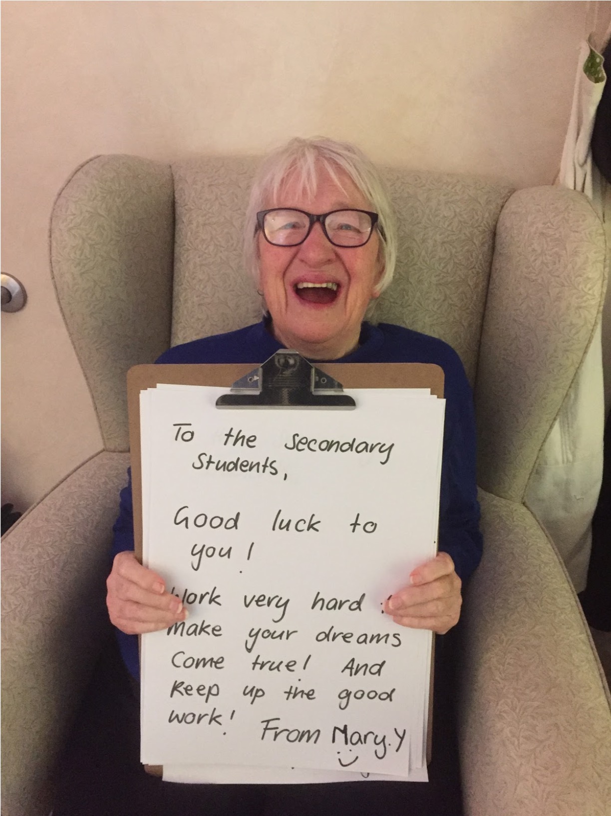 Mary, a resident is sitting in an armchair laughing at the camera. She is holding a large clipboard with a hand written message attached. The message reads: "To the Secondary Students, Good luck to you! Work very hard and make your dreams come true! And keep up the good work! From Mary Y"