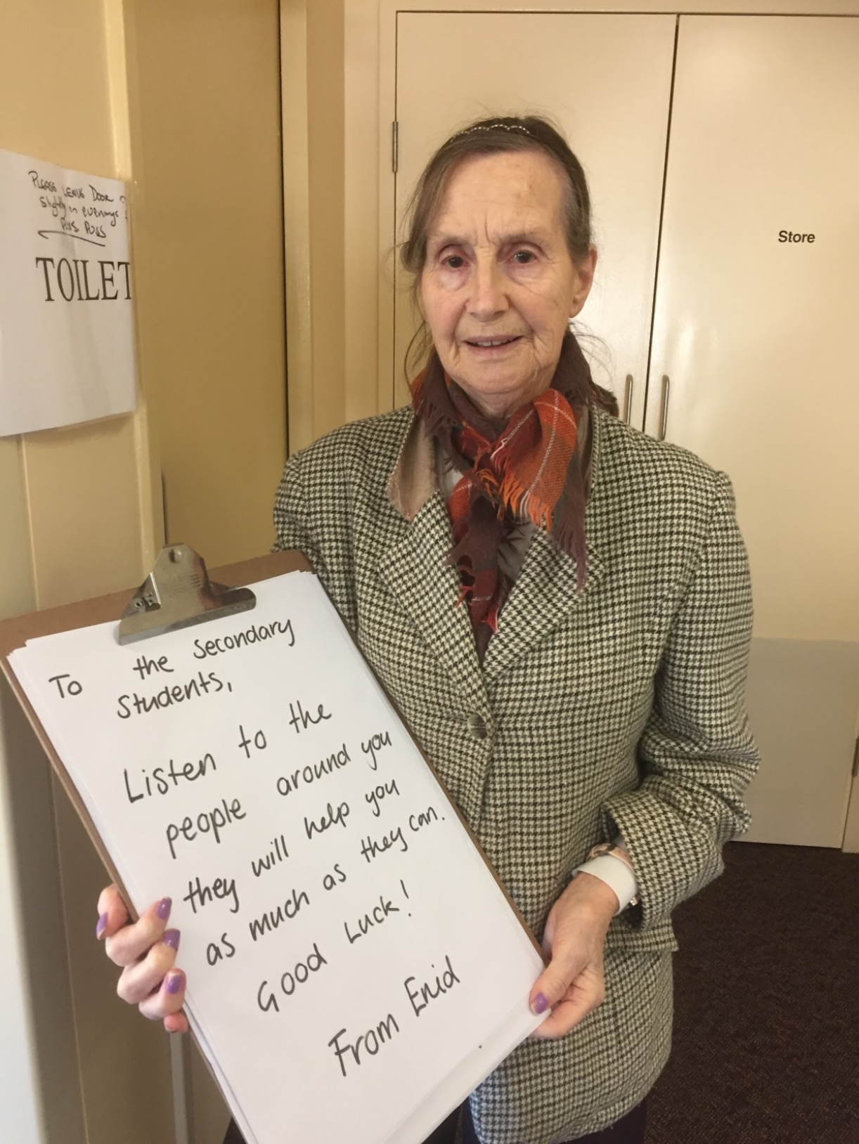 Enid, a resident, holds up a large clipboard with a hand written message which reads: "To the Secondary Students, Listen to the people around you, they will help you as much as they can. Good luck! From Enid."