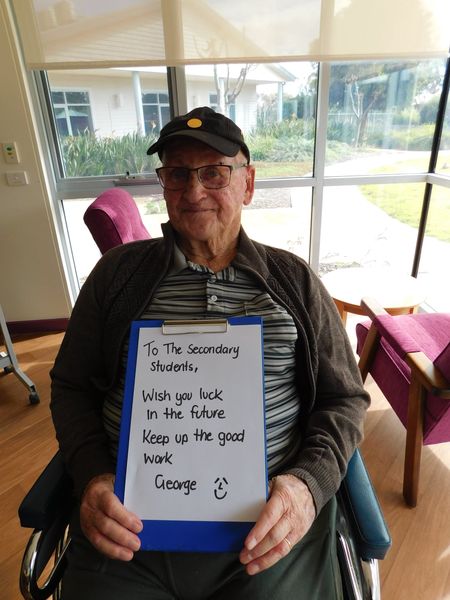 George a resident, sits in a chair holding a blue clipboard with a hand written message on it which reads "To the Secondary Students, Wish you luck in the future. Keep up the good work. George."