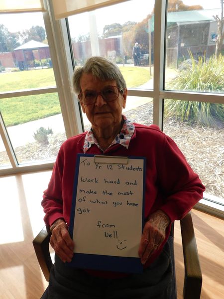 Nell, a resident, is sitting in a chair holding up a clipboard with a hand written message on it which reads "To Year 12 Students, Work hard and make the most of what you have got. From Nell"