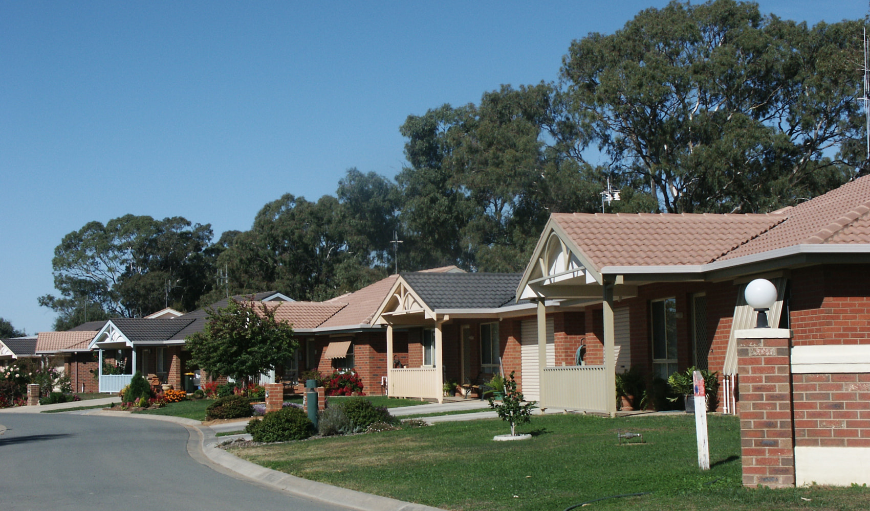 A outside view of the village at Gwandalan Court showing the red brick houses and their front lawns. It's a sunny day and the village is surrounded by tall gum trees. 