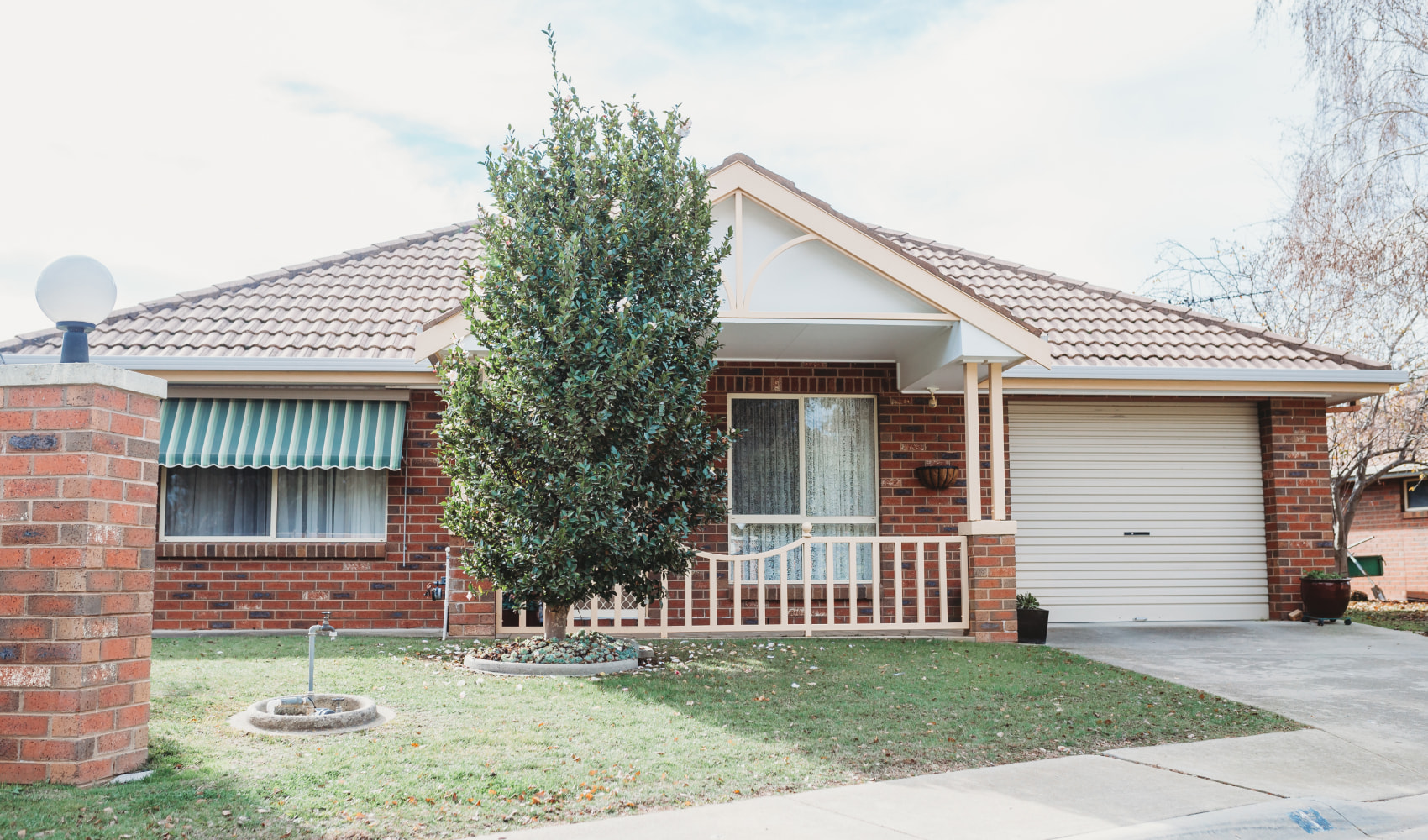 One of the single level red brick retirement houses at Gwandalan Court. The front garden shows a green lawn and a small leafy tree. 