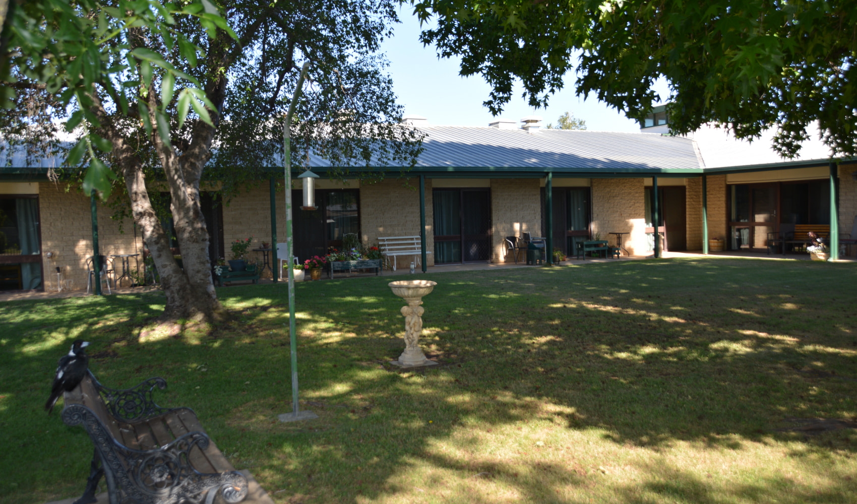 A view of the pretty dappled light in the garden at Numurkah Pioneer's Memorial Lodge showing large trees, a park bench and a bird bath. 