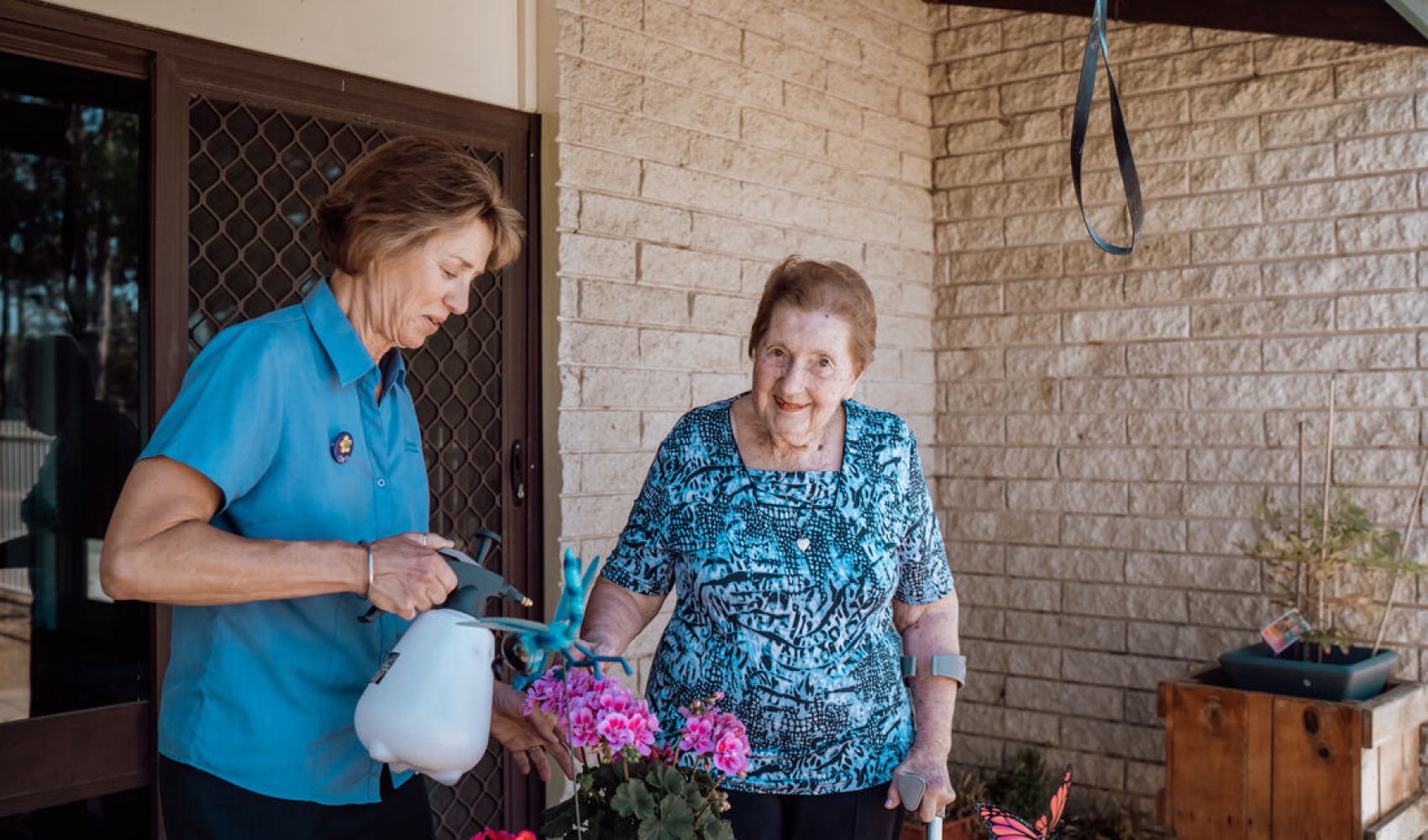 A female staff member and female resident watering pink potted flowers. The resident is smiling at the camera. 