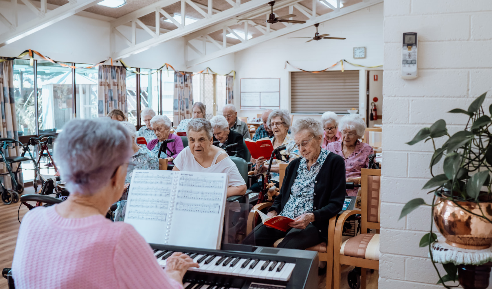 A large group of residents sing in a choir, whilst a lady plays the keyboard. They are all seated in a large bright airy room. 