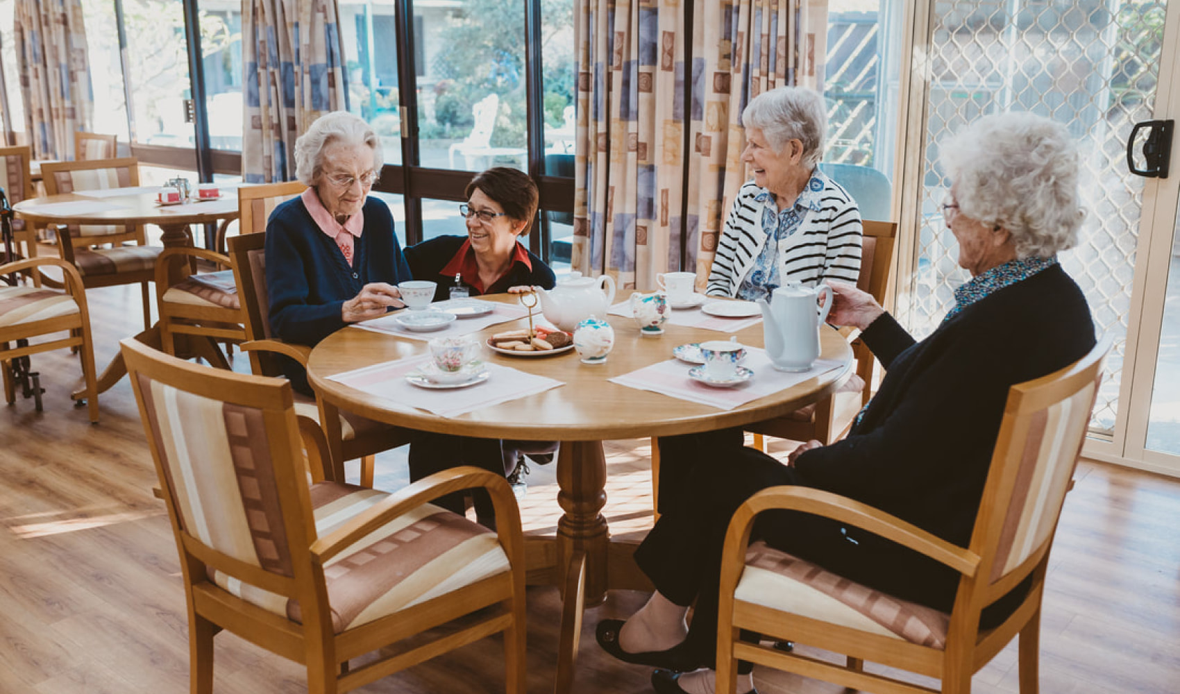 3 female residents and a female staff member are happily enjoying afternoon tea together around a round table. They are seated in an airy room with lots of sunlight. The women are laughing. 