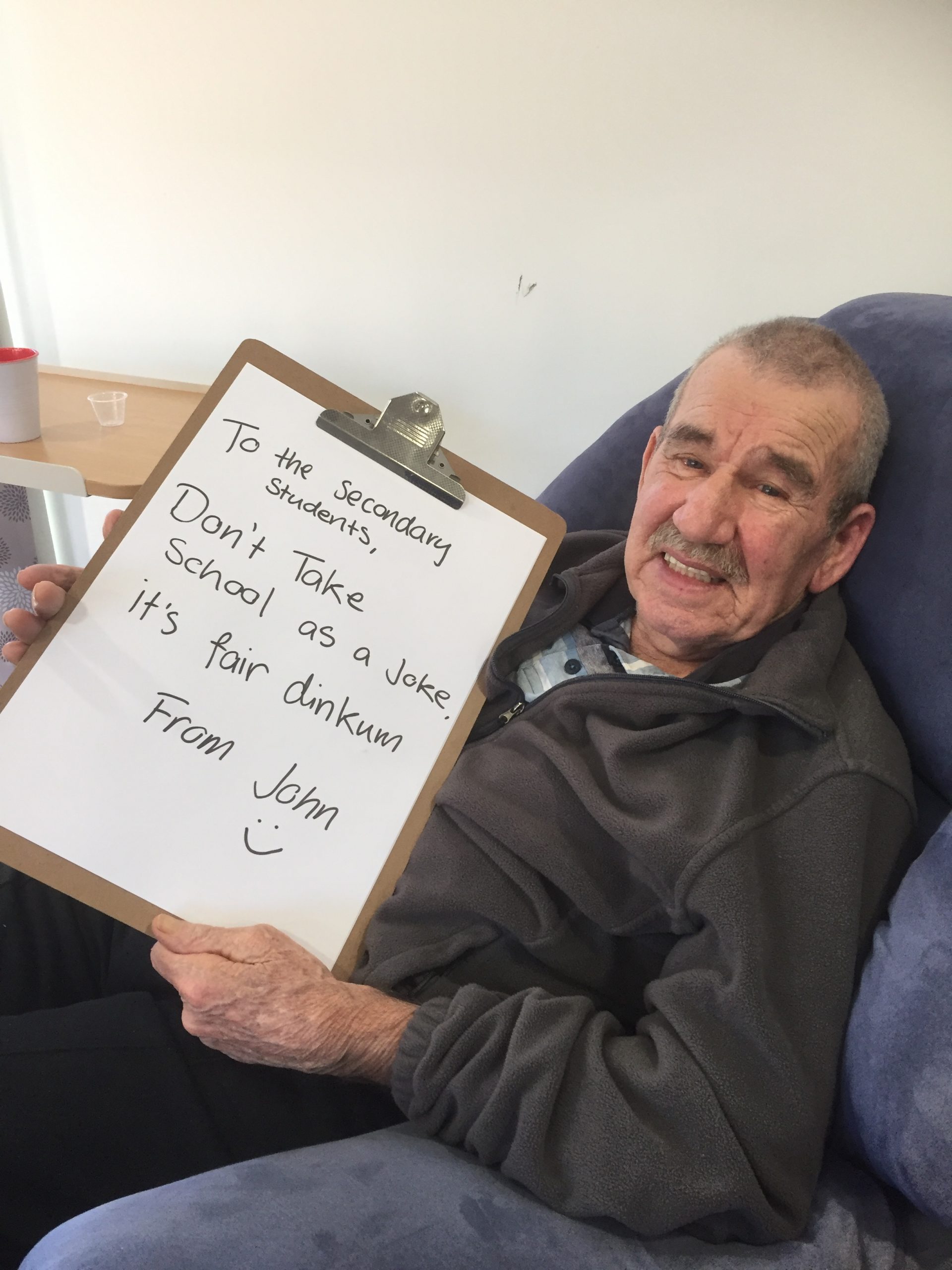 A male resident holds up a hand written sign on a clipboard which reads: "To the secondary students, Don't take school as a joke. It's fair dinkum. From John"
