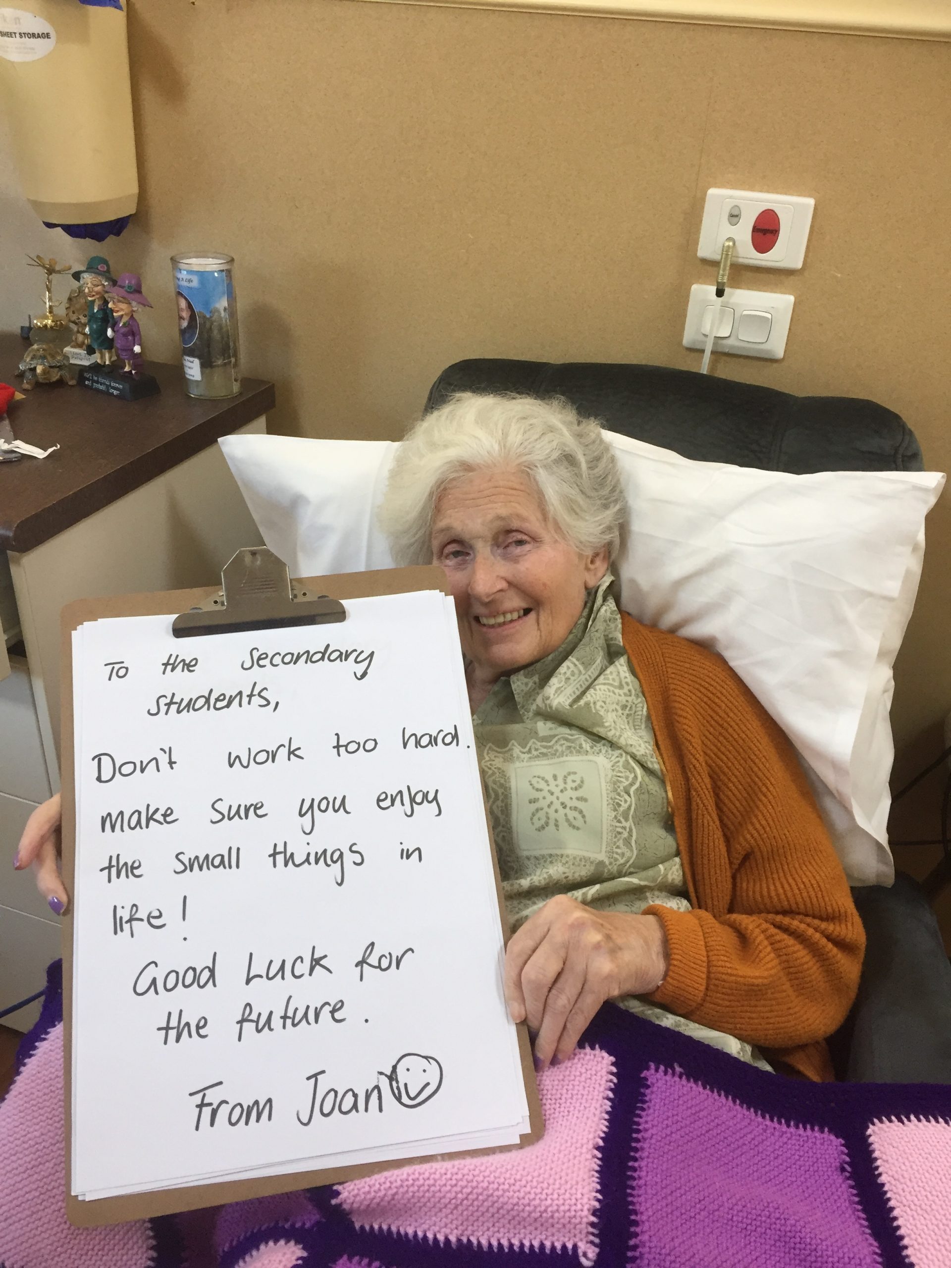 A female resident smiles at the camera whilst holding up a large clipboard with a hand written message to Secondary Students, which reads: "To the Secondary Students, Don't work too hard. Make sure you enjoy the small things in life. Good luck for the future. From Joan." 