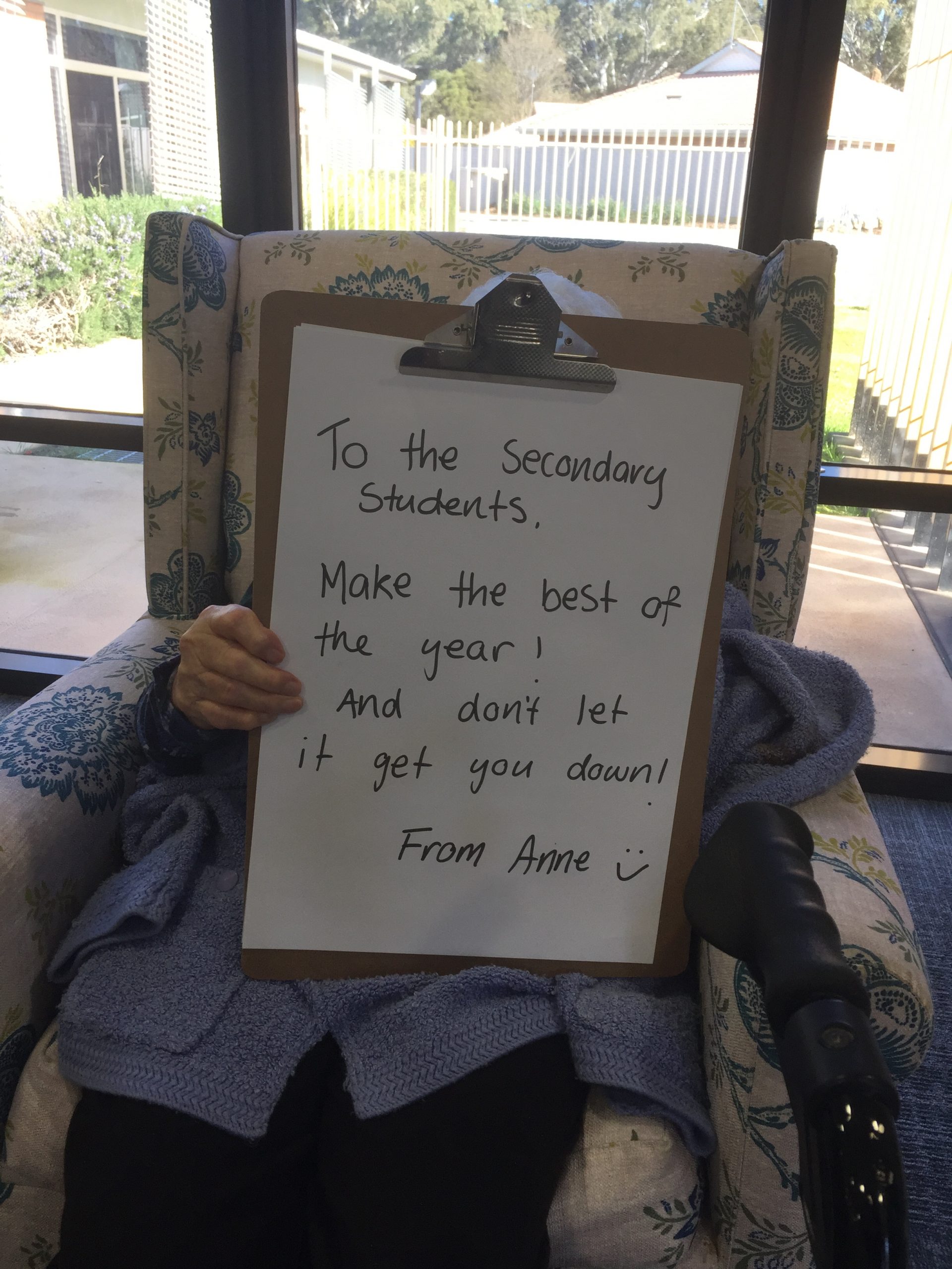 Anne, a resident, sits in an armchair and holds up a clipboard with a hand written message on it. The clipboard is obscuring her face. The message reads: "To the Secondary Students, Make the best of the year! And don't let it get you down! From Anne"