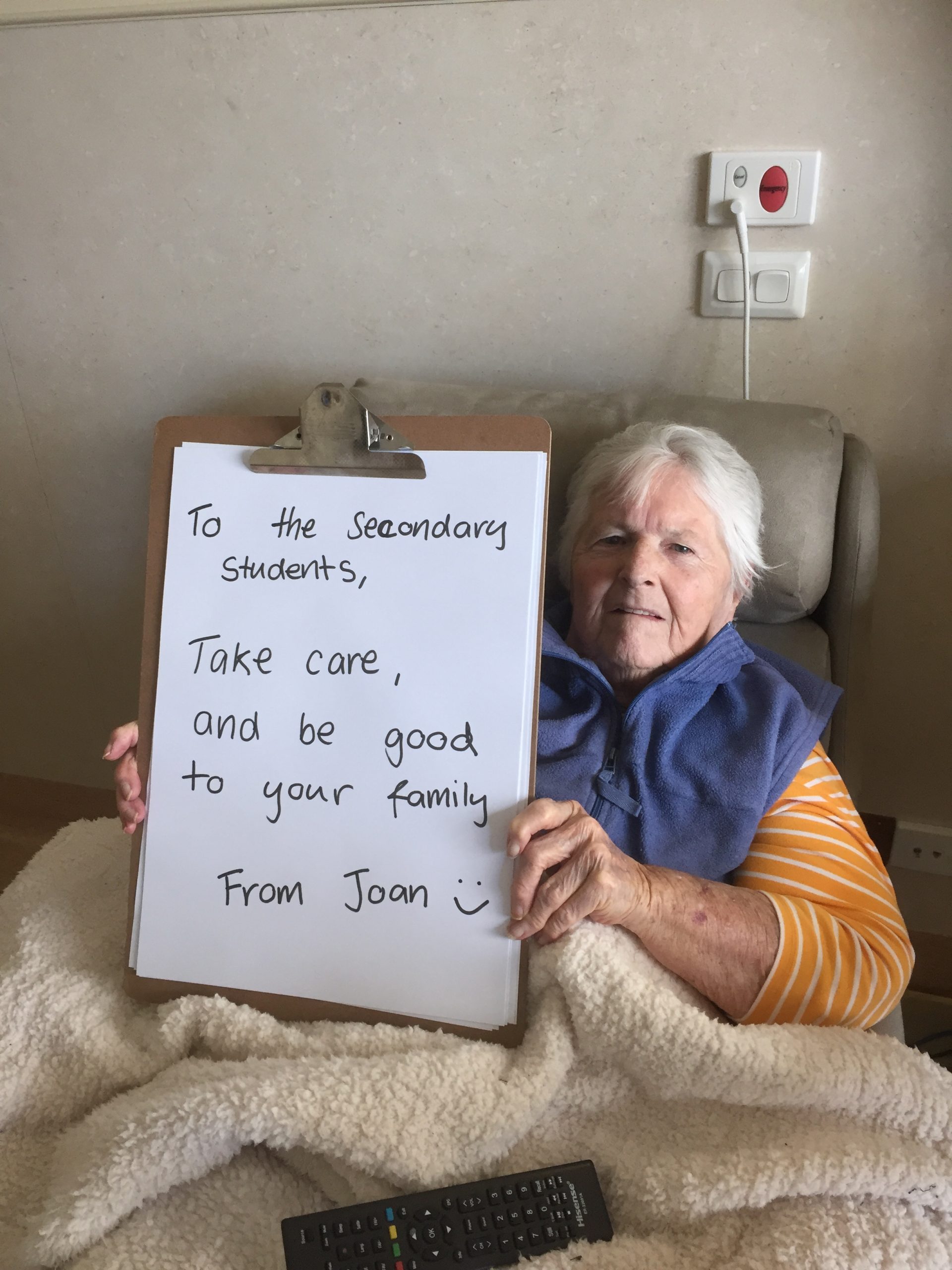 Joan sits in an armchair holding up a large clipboard with a hand written message on it which reads "To the Secondary Students, Take care, and be good to your family. From Joan"