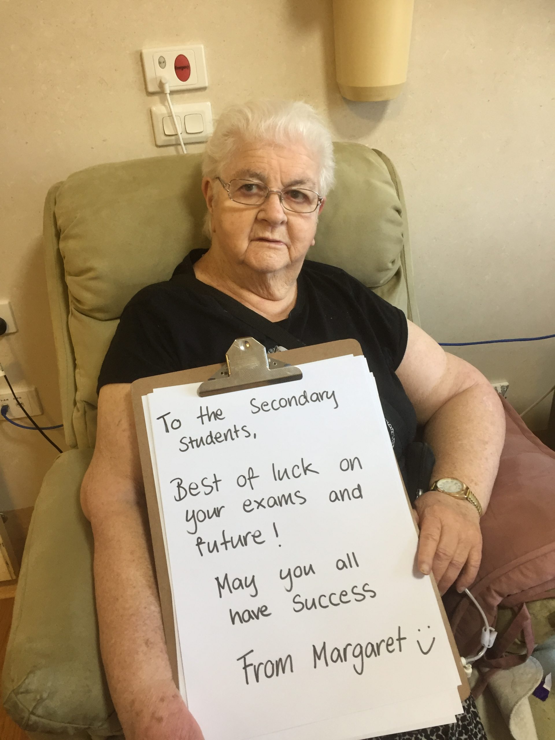 Margaret, a resident sits in an armchair holding a large clipboard with a hand written message which reads "To the Secondary Students, Best of luck on your exams and future! May you all have success. From Margaret."