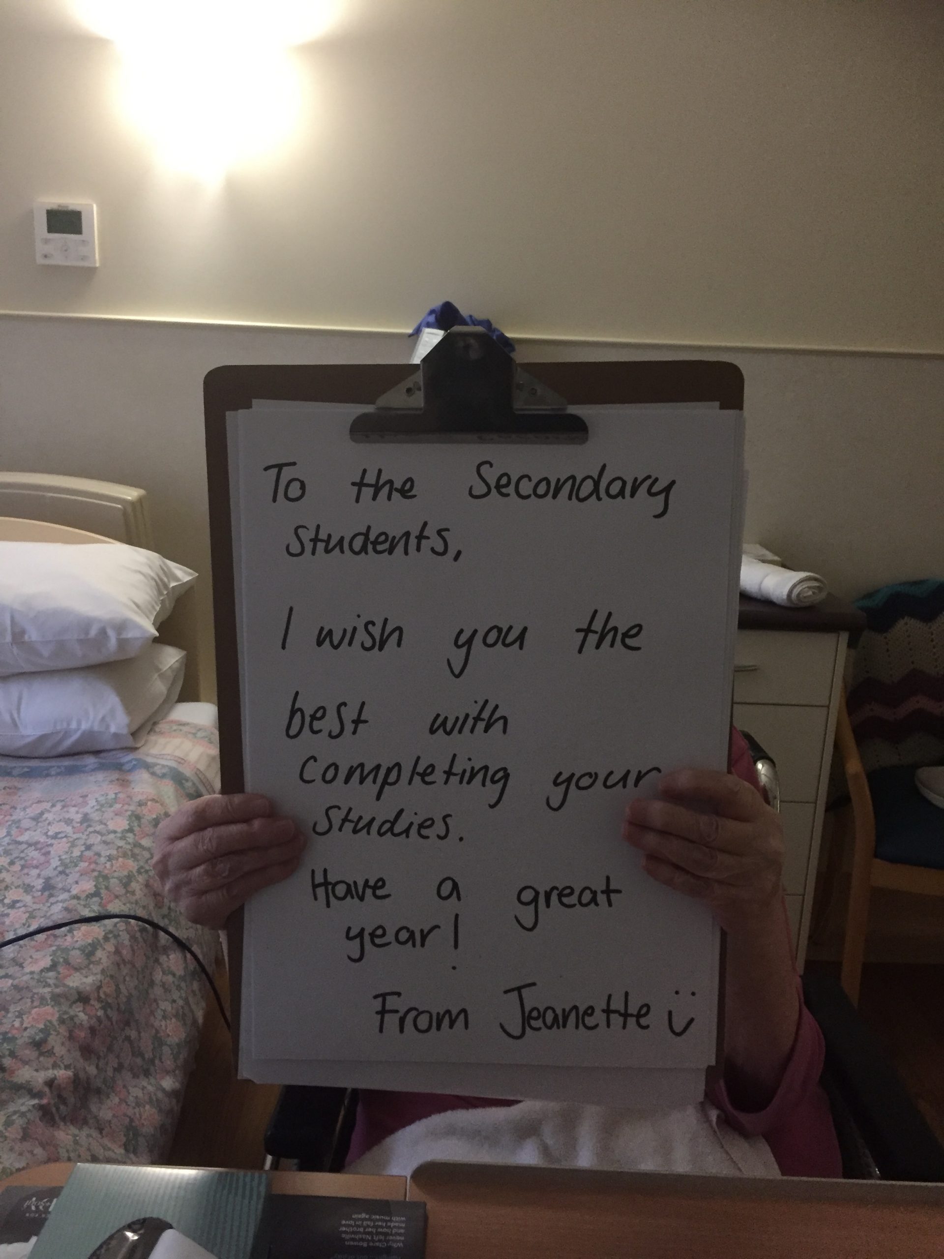 Jeanette, a resident, is obscured behind a large clipboard she is holding, which has a personal message written on it which reads "To the Secondary Students, I wish you the best with completing your studies. Have a great year! From Jeanette"
