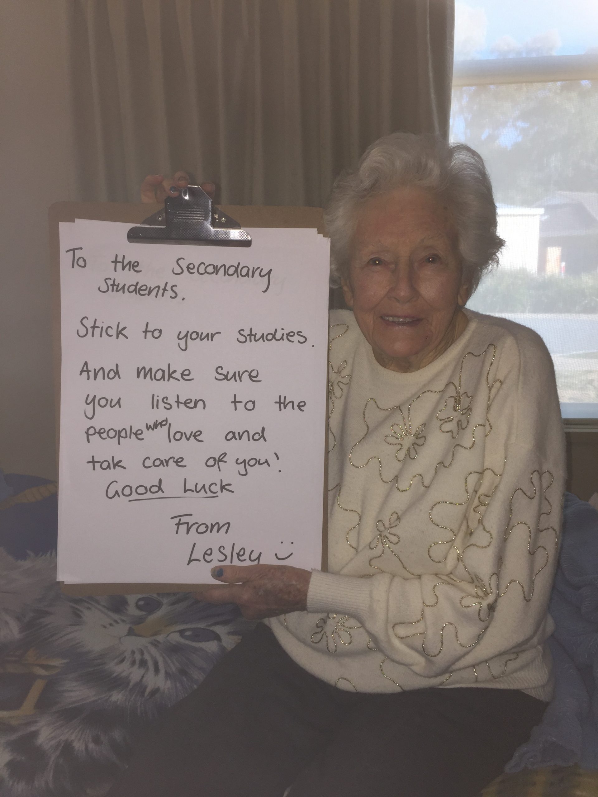 Lesley sits on the edge of a bed holding up a large clipboard which reads "To the Secondary Students, Stick to your studies. And make sure you listen to the people who love you and take care of you! Good lucky. From Lesley"