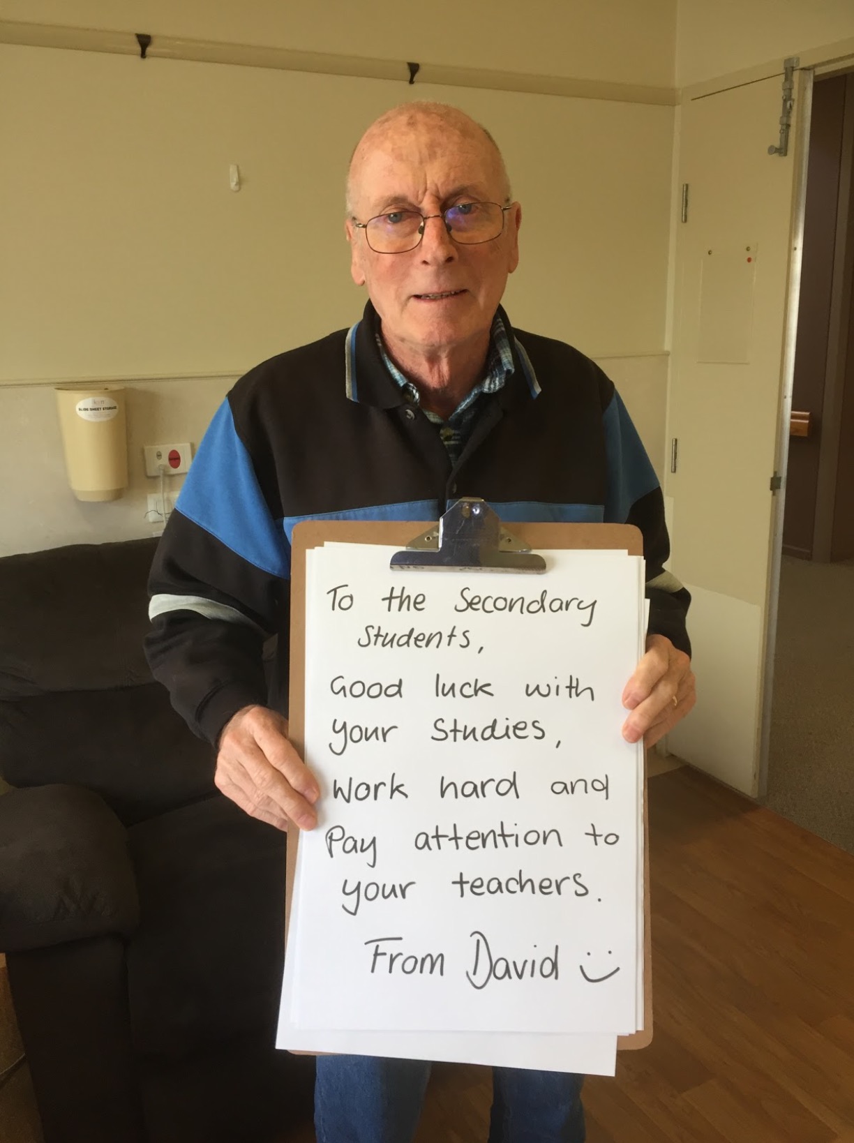 David, a resident, is standing inside holding up a large clipboard with a hand written message on it which reads, "To the Secondary Students, Good luck with your studies. Work hard and pay attention to your teachers. From David."